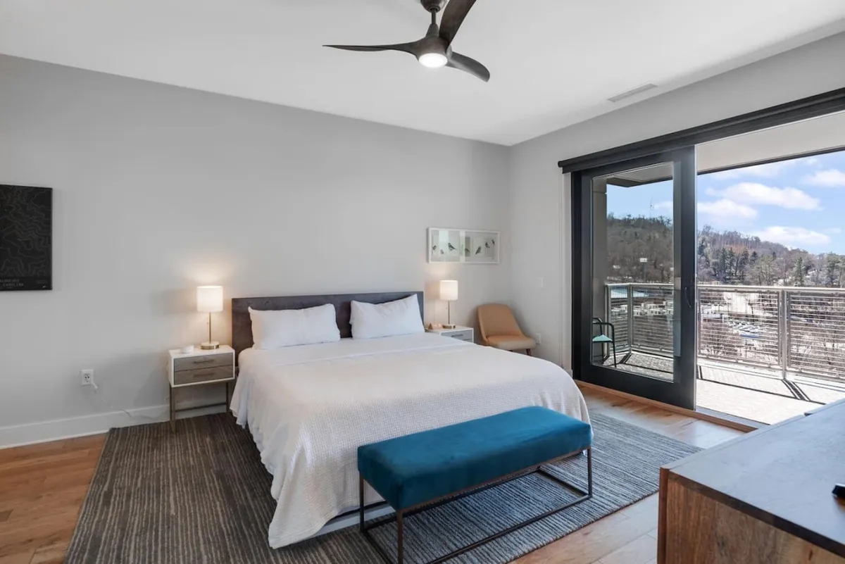 A tranquil king bedroom featuring a plush king-size bed with luxurious bedding, nightstands with lamps, a modern ceiling fan, and direct access to a balcony boasting a scenic view. The room is bathed in soft natural light, creating a peaceful, serene retreat ideal for rest and privacy.
