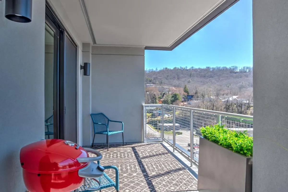 A casual balcony setup featuring a red grill, blue chairs, and a green planter, all oriented toward a balcony view. The atmosphere is inviting and relaxed, ideal for enjoying morning coffee or a sunset barbecue while taking in the scenery.
