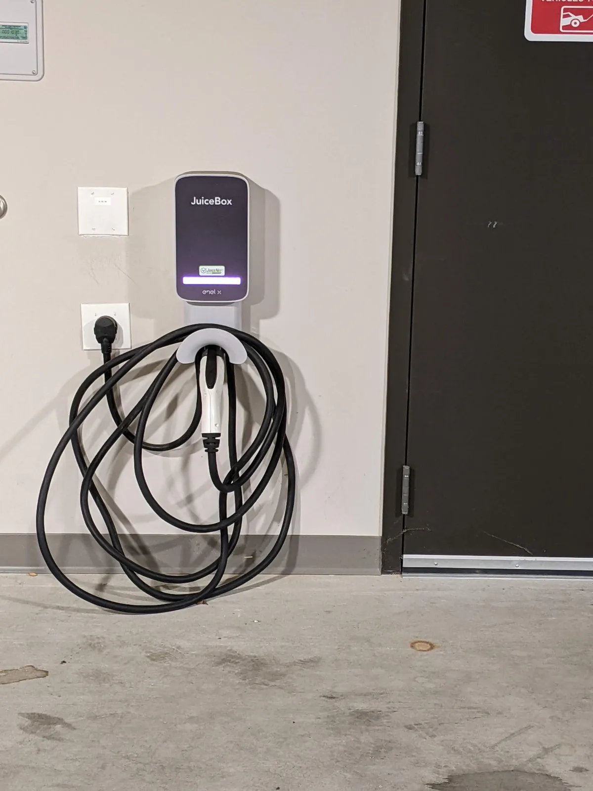 A modern garage setting featuring a JuiceBox electric vehicle charger mounted on the wall, a nearby power outlet, and a doorway leading to the interior. The space is clean, contemporary, and signals eco-friendly convenience.