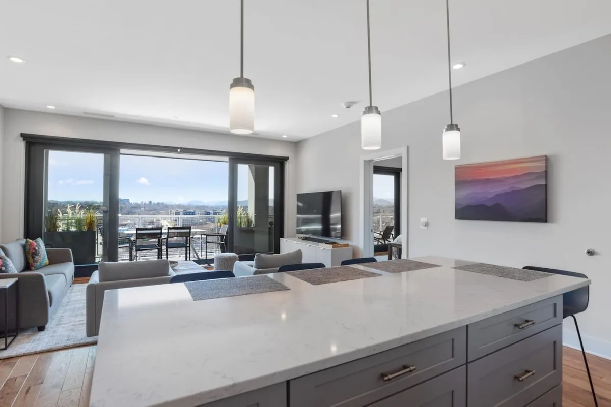 This image captures the open-plan living area with a kitchen island, large windows that let in abundant natural light, a comfortable seating area, and a clear view of the mountains. Artwork on the wall adds a touch of personality and warmth to the space, while the vantage point allows viewers to see how the living, dining, and kitchen areas flow together.