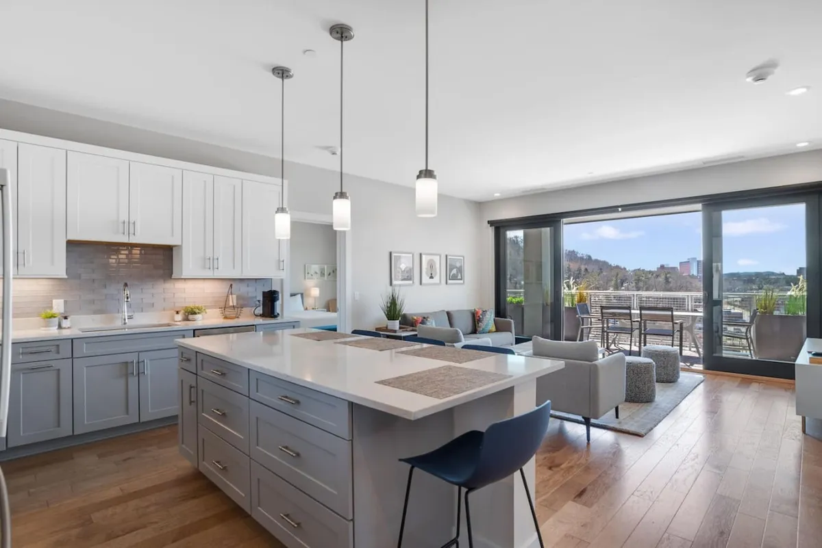 A welcoming view of the kitchen with a prominent island countertop, modern appliances, and a seamless transition to the adjacent seating area. Large windows frame the backdrop, flooding the space with natural light and providing glimpses of the scenery outside.