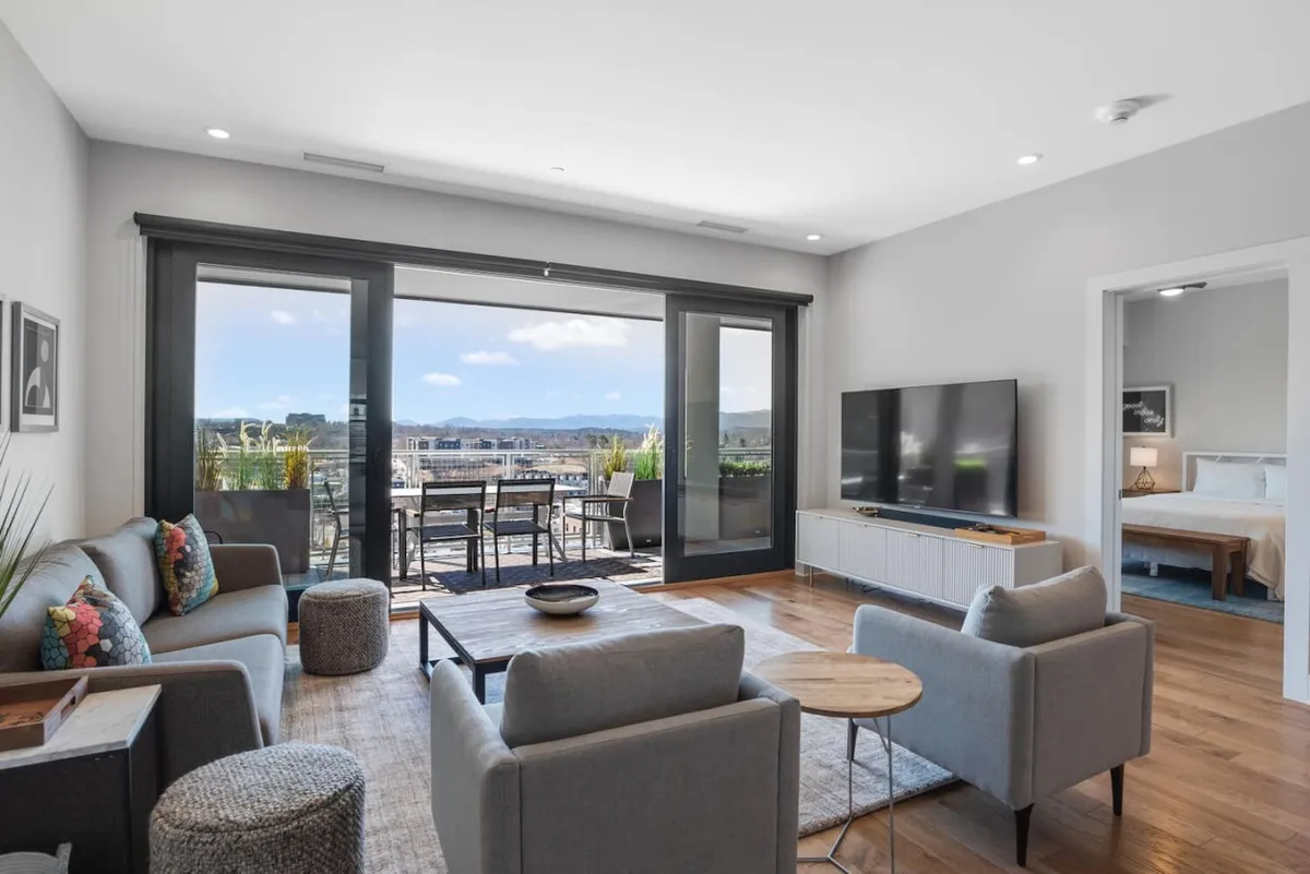 The image showcases a bright, modern living area with plush seating, floor-to-ceiling windows framing sweeping mountain views, and direct access to a balcony set for dining. The welcoming space seamlessly blends indoor comfort with outdoor beauty, highlighting the condo’s contemporary style and the awe-inspiring Asheville landscape.