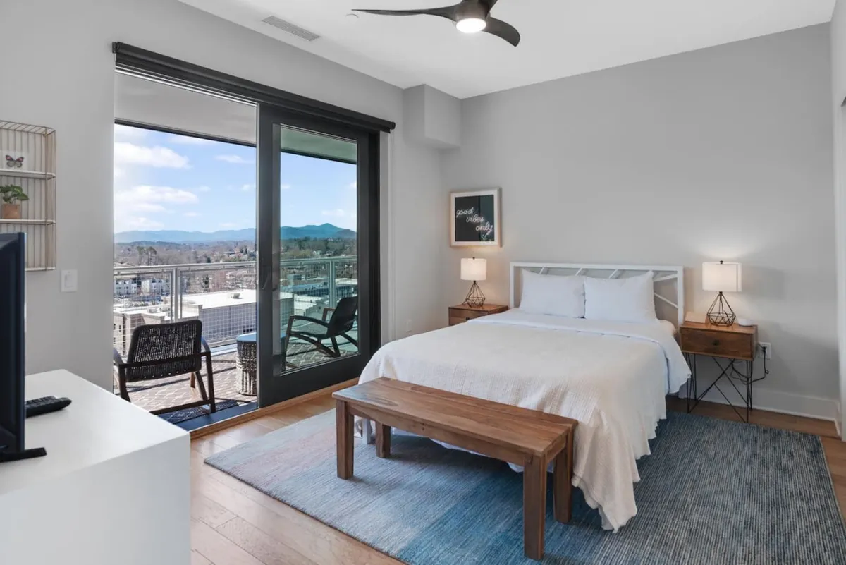 A serene queen bedroom with a comfortable bed, nightstand and lamp, wooden bench at the foot of the bed, TV, and a balcony view. The soft lighting and elegant decor enhance the peaceful, inviting atmosphere.
